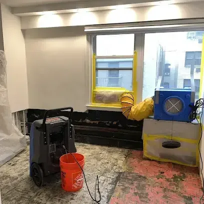 Dehumidifier and blue air mover drying a room with the flooring torn out and yellow duct by the window