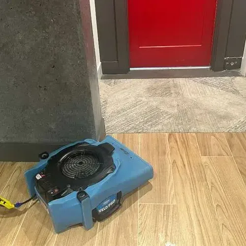 Air mover aimed across a wood-look tile floor beside an open doorway during a dry-out