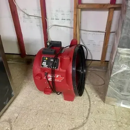 Red air mover on a tile floor beside a wall opened to the studs and plastic sheeting