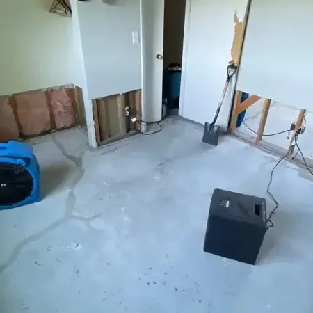 Blue air mover and a dehumidifier on a bare floor beside walls with the lower drywall cut out